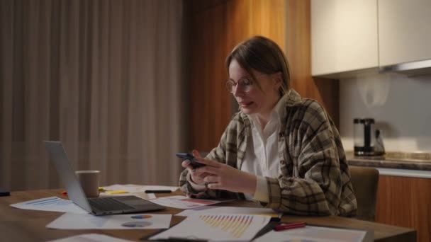 Millennial woman employee sit by work desk chat in phone messenger app taking break in computer job. Young lady worker manager browse web in mobile application on smartphone, office from home — Stockvideo