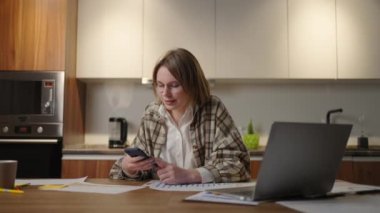 Genç bayan evde çalışıyor. Dizüstü bilgisayarda yazmak ve telefondaki mesajlara cevap vermek. Akıllı telefon kullanan ve odada dinlenen bir kadın.