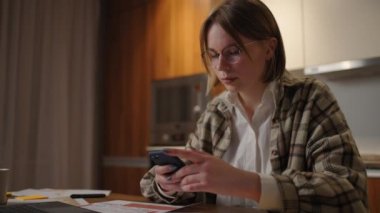 Genç bayan evde çalışıyor. Dizüstü bilgisayarda yazmak ve telefondaki mesajlara cevap vermek. Akıllı telefon kullanan ve odada dinlenen bir kadın.