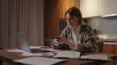 Evde dizüstü bilgisayarla çalışırken cep telefonu kullanan mutlu bir kadın. Gözlüklü, akıllı telefonlu, gülümseyen, havalı bir kadın. Cep telefonundaki güzel şık bayan arama sitesi