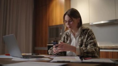 Evde dizüstü bilgisayarla çalışırken cep telefonu kullanan mutlu bir kadın. Gözlüklü, akıllı telefonlu, gülümseyen, havalı bir kadın. Cep telefonundaki güzel şık bayan arama sitesi