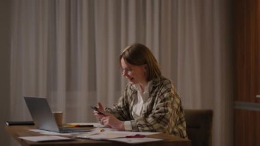 Evde dizüstü bilgisayarla çalışırken cep telefonu kullanan mutlu bir kadın. Gözlüklü, akıllı telefonlu, gülümseyen, havalı bir kadın. Cep telefonundaki güzel şık bayan arama sitesi