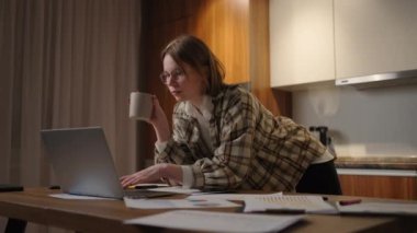 Oturma odasında oturmuş kahve içerken dizüstü bilgisayarda çalışan güzel genç bir kadın. Dizüstü bilgisayarda çalışan mutlu, sıradan bir kadın. Evde dizüstü bilgisayar ve belgelerle çalışmak