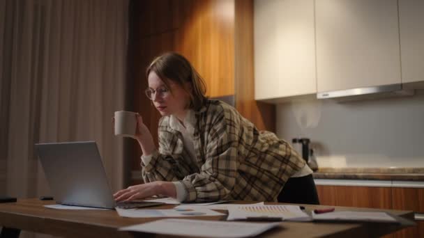 한 젊은 아름다운 사업가가 커피를 마시고 집에서 일하다가 탁자 위에 서 있는 노트북 화면을 들여다보고 있습니다. 우편물 과 커피를 본다. 집에서 멀리 떨어져 일하는 모습 — 비디오