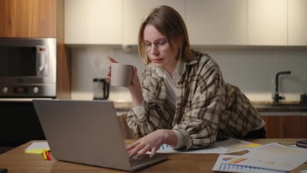 Uma jovem mulher de negócios bonita trabalha em casa com uma xícara de café e olha para uma tela de laptop em pé na mesa. Ver e-mail e beber café. Trabalho remoto em casa — Vídeo de Stock