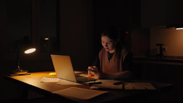 Красивая женщина работает допоздна дома, используя ноутбук и получая текстовые сообщения на смартфоне. Руки женщины, отправляющей смс на мобильный смартфон для общения и чата в соцсети — стоковое видео