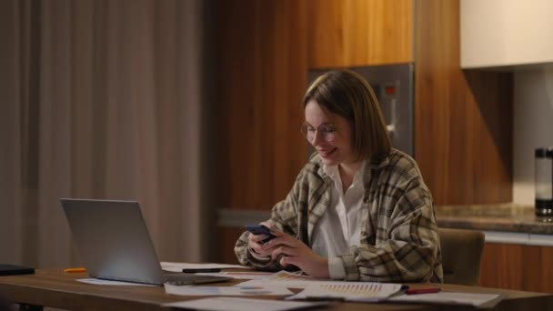 Mujer feliz usando el teléfono móvil mientras trabaja en casa con el ordenador portátil. Mujer guay sonriente con gafas de mensajería con teléfono inteligente. Hermosa señora elegante sitio de navegación en el teléfono celular — Vídeos de Stock