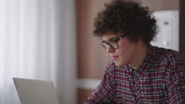 Ciddi görünümlü bir kıvırcık adam, modern bir mutfakta oturan bir dizüstü bilgisayarda çalışıyor. Genç serbest çalışan öğrenci bilgisayarını kullanıyor. İnternetten çalışıyor.