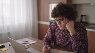 Düşünceli, ciddi, kıvırcık, serbest çalışan bir adam rahat bir ofis odasında oturuyor dizüstü bilgisayarla çalışıyor, iş meselelerini enine boyuna düşünüyor ve telekomünikasyon işi yapıyor.