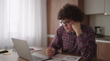 Düşünceli, ciddi, kıvırcık, serbest çalışan bir adam rahat bir ofis odasında oturuyor dizüstü bilgisayarla çalışıyor, iş meselelerini enine boyuna düşünüyor ve telekomünikasyon işi yapıyor.