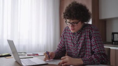 Kıvırcık saçlı, gözlüklü çekici erkek öğrenci evde çalışıyor, dizüstü bilgisayar kullanıyor, not defteri yazıyor. Kolej öğrencisi dizüstü bilgisayarı kullanarak online öğrenme semineri yapıyor.
