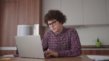Ciddi görünümlü bir kıvırcık adam, modern bir mutfakta oturan bir dizüstü bilgisayarda çalışıyor. Genç serbest çalışan öğrenci bilgisayarını kullanıyor. İnternetten çalışıyor.