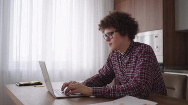 Ciddi görünümlü bir kıvırcık adam, modern bir mutfakta oturan bir dizüstü bilgisayarda çalışıyor. Genç serbest çalışan öğrenci bilgisayarını kullanıyor. İnternetten çalışıyor.