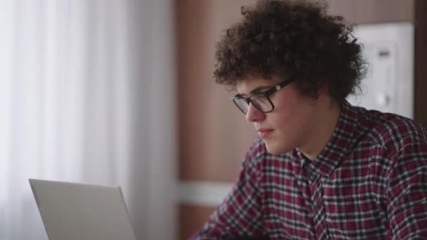 Ciddi görünümlü bir kıvırcık adam, modern bir mutfakta oturan bir dizüstü bilgisayarda çalışıyor. Genç serbest çalışan öğrenci bilgisayarını kullanıyor. İnternetten çalışıyor. — Stok video
