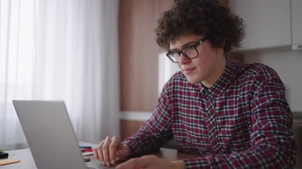 Een Curly man met een serieuze blik werkt op een laptop, zittend in een moderne keuken. Jongeman freelancer student met behulp van laptop studeren online werken vanuit huis in internet — Stockvideo