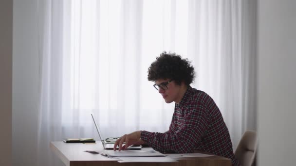 Um homem encaracolado com um olhar sério trabalha em um laptop, sentado em uma cozinha moderna. Jovem estudante freelancer usando laptop estudando on-line trabalhando em casa na internet — Vídeo de Stock