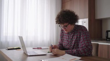 Kıvırcık saçlı, gözlüklü çekici erkek öğrenci evde çalışıyor, dizüstü bilgisayar kullanıyor, not defteri yazıyor. Kolej öğrencisi dizüstü bilgisayarı kullanarak online öğrenme semineri yapıyor.