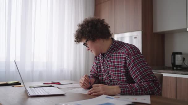 Göndör hajú férfi diák vonzó fiatal fiú szemüvegben otthon tanul laptoppal gépel, jegyzetfüzetbe ír. Egyetemista használó laptop számítógép távoktatási online szeminárium — Stock videók
