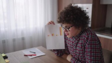 Kıvırcık saçlı, gözlüklü, ofis masasında oturan, kameraya bakan ve grafiksel form grafiği olan bir tableti işaret eden bir iş adamı.