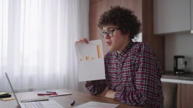 Kıvırcık saçlı, gözlüklü, ofis masasında oturan, kameraya bakan ve grafiksel form grafiği olan bir tableti işaret eden bir iş adamı.