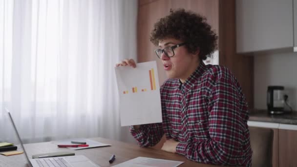 Lockig - mit Brille Geschäftsmann sitzt im Büro von zu Hause aus Schreibtisch Blick auf Kamera und zeigt auf ein Tablet mit Finanzinformationen in grafischer Form Spaltendiagramm — Stockvideo