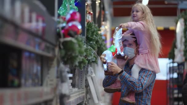 Μπαμπάς και κόρη επιλέγουν διακοσμήσεις και γιρλάντες με παιχνίδια για χριστουγεννιάτικα δέντρα και στο σπίτι στο κατάστημα. Χριστουγεννιάτικες γιρλάντες και διακόσμηση — Αρχείο Βίντεο