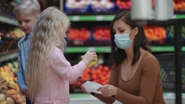 Мати в захисній масці з двома дітьми купує продукти в супермаркеті. Купівля їжі овочів і фруктів з дітьми — стокове відео