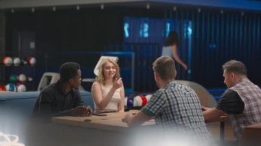 A multi-ethnic group of friends in a bowling club are chatting at the table and smiling, rejoicing and high-fiving a friend who knocked out a strike. Black man and woman