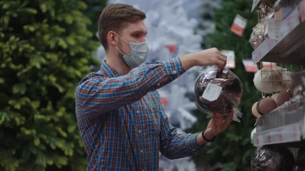 Compras en pandemia y cuarentena. Un hombre con una máscara protectora en una joyería y guirnaldas con juguetes para árboles de Navidad y en casa. Guirnaldas de Navidad y decoración. — Vídeos de Stock