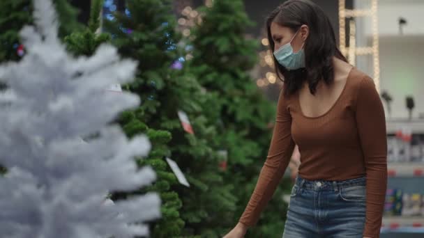 Una mujer con una máscara protectora en una joyería y guirnaldas con juguetes para árboles de Navidad y en casa. Guirnaldas de Navidad y decoración — Vídeos de Stock