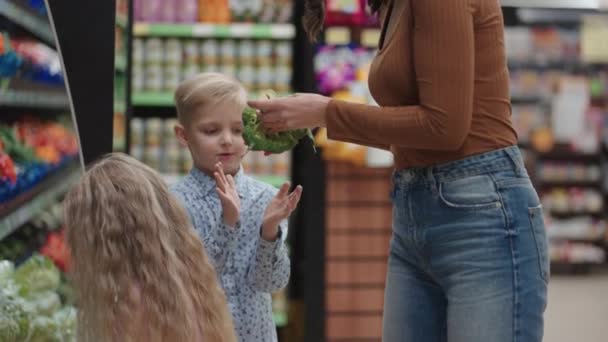 一位戴着防护面具的母亲带着两个孩子在超级市场买食品杂货。与儿童一起购买食品、蔬菜和水果 — 图库视频影像