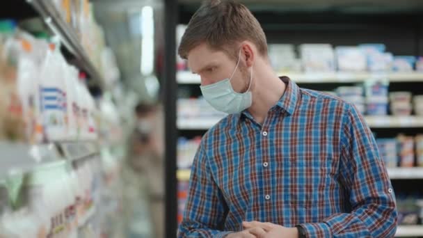 A young man in a supermarket in a protective mask chooses milk and chilled foods — ストック動画