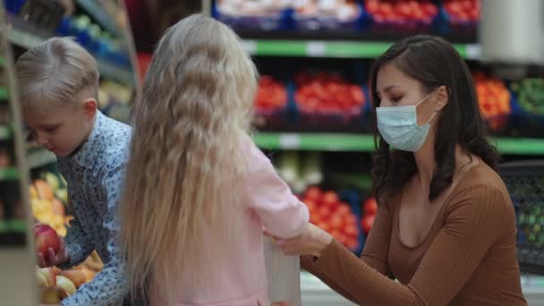 2人の子供を持つ保護マスクの母親は、スーパーで食料品を購入しています。子供と一緒に野菜や果物を買う — ストック動画