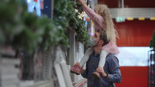 Vater und Tochter, die in einem Einkaufszentrum auf ihren Schultern sitzen, wählen während der Pandemie in Zeitlupe einen Kranz für das Haus und den Weihnachtsbaum für Weihnachten. — Stockvideo