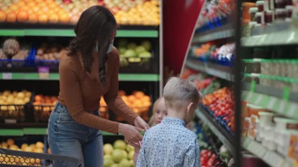 2人の子供を持つ保護マスクの母親は、スーパーで食料品を購入しています。子供と一緒に野菜や果物を買う — ストック動画