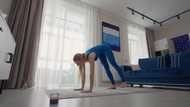 Mujer joven está practicando yoga en casa relajándose en posición corporal sencilla sentada en el suelo con hermosos muebles alrededor. mujer delgada en ropa deportiva de pie sobre las rodillas, inclinándose hacia atrás y haciendo pose de camello — Vídeos de Stock