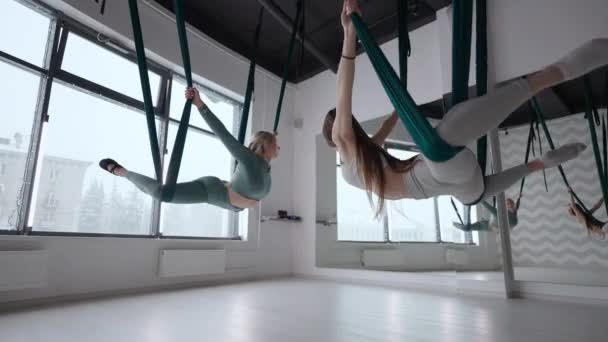 Entrenadora y joven practicante de gimnasia aérea con suspensiones en el gimnasio. Grupo de dos jóvenes hermosas mujeres yoguis haciendo práctica de yoga aéreo en hamacas verdes en el gimnasio — Vídeo de stock