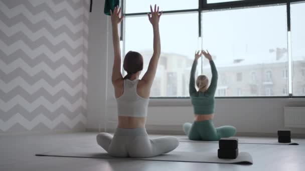 Eine Gruppe junger Frauen dehnt sich in einer Turnhalle mit Fenstern. Zwei attraktive Sportmädchen trainieren Nauka Asana Boot Yoga Pose auf schwarzer Matte im Fitnessunterricht. Zwei Frauen strecken im Yoga-Studio die Beine zusammen — Stockvideo