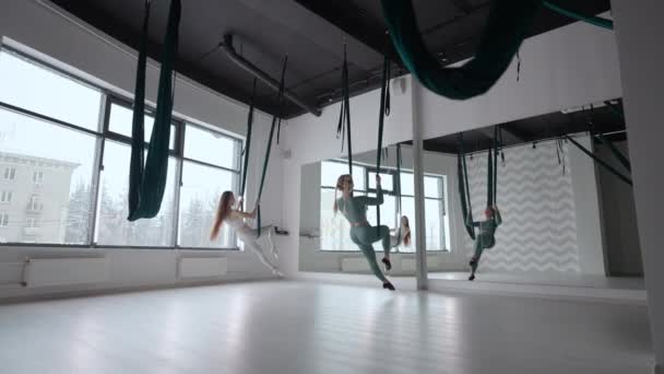 Deux jeunes femmes yogi qui pratiquent le yoga aérien dans des hamacs verts dans un club de fitness. Belles femmes travaillant en classe, effectuant de l'aéro yoga. Variation de Parsvottanasana, pose pyramidale — Video