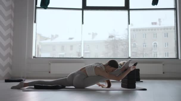 Grupa młodych kobiet rozciągających się w siłowni z oknami. Dwie atrakcyjne sportowe dziewczyny ćwiczą naukę asana boat yoga pose na czarnej macie w klasie fitness. Dwie kobiety rozciągające nogi razem w studiu jogi — Wideo stockowe