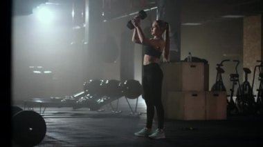 Güçlü bir kadın çaba sarf eder ve zorlukların üstesinden gelir. Karanlık bir spor salonunda bir dambılı kaldırır. Spor salonunda spor salonundaki ağırlık kaldıran kadın..