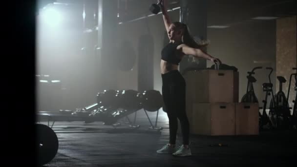 Une femme forte fait des efforts et surmonter les difficultés soulève un haltère dans un gymnase sombre. Fitness femme haltères haltères haltères haltères entraînement dans le club de gym. — Video