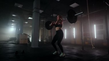 Vücut geliştiren bir kadın spor salonunda ağırlık çalışıyor. Bir kadın halterci karanlık bir spor salonunda halter kaldırır. Kadın Vücut Geliştirici Hardcore Eğitiminde Ağırlık Kaldırma Egzersizleri Yapıyor