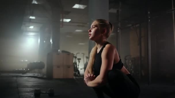 Eine junge schlanke Frau bereitet sich vor dem Training vor und wärmt sich auf. Trampen und Dehnen der Muskeln nach einem anstrengenden harten Training im dunklen Innenraum des Fitnessraums — Stockvideo