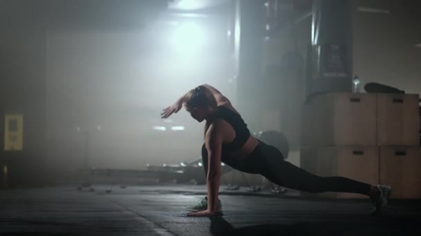 Een jonge slanke vrouw bereidt zich voor en warmt zich op voor de training. Hitching en stretching spieren na een vervelende harde training in het donkere interieur van de fitnessruimte — Stockvideo