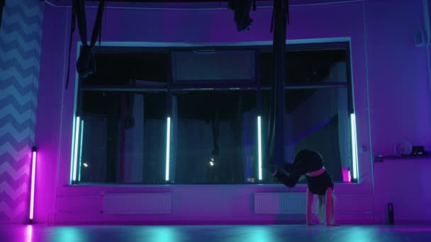A woman does yoga and stretching on a hanging hammock in neon light. Young pretty slim body fitness girl practicing fly yoga in the gym. — Stock Video