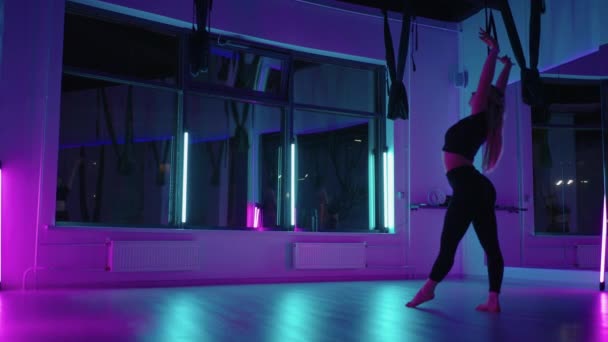 Bir kadın yoga yapıyor ve neon ışıkta asılı bir hamakta geriniyor. Bir kadın stüdyoda hamakta uçuyor, esneme hareketleri yapıyor ve vücut egzersizleri yapıyor. Havadaki egzersizler. — Stok video