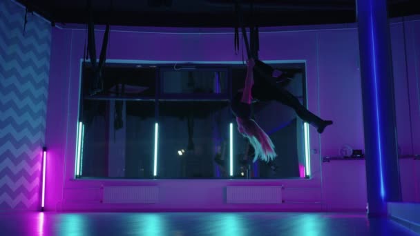 Una donna fa yoga e si estende su un'amaca appesa in luce al neon. Una donna sta volando in un hammock nello studio facendo lo stretching e gli esercizi del corpo. Esercizi in aria — Video Stock