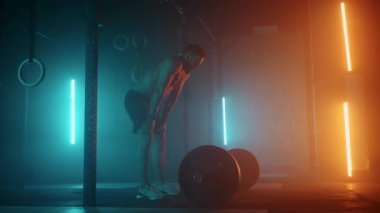 Çıplak gövdeli tişörtü olmayan güçlü bir adam halteri başının üzerine kaldırıyor ağır çekimde renkli ışıkla ağır çekimde bir halter. Spor salonunda mavi turuncu neon ışık