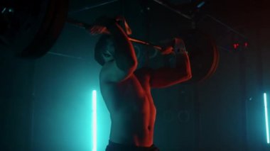 Mavi-turuncu neon ışıklı erkek kaldırıcı spor salonunda antrenmana hazırlanıyor. Çıplak gövdeli güçlü bir adam halteri başının üzerine kaldırır. Ağırlık kaldırma egzersizi yapar..